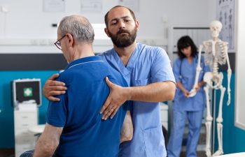 Orthopedic nurse stretching spinal cord and shoulder of elder patient with injury for physiotherapy. Chiropractor giving help to senior man with back pain for physical recovery.